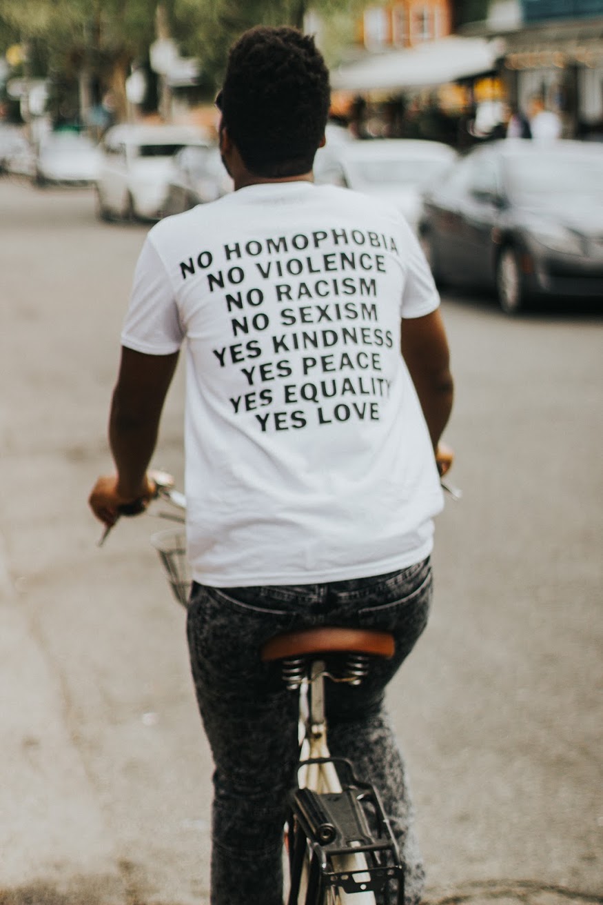 A riding a bike. The photo is focus on the back of the white shirt with black writing. The text has our signature statement:
No Homophobia
No Violence
No Racism
No Sexism
Yes Kindness
Yes Peace
Yes Equality
Yes Love
