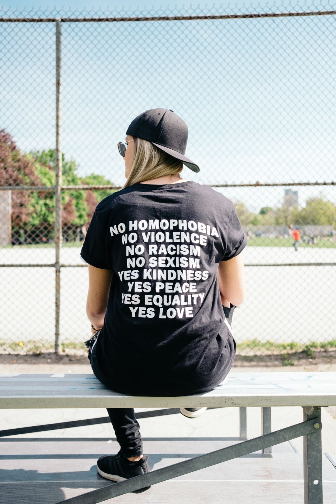 A person sitting on a bench the photo is focus on the back of the black shirt with white writing. The text has our signature statement:
No Homophobia
No Violence
No Racism
No Sexism
Yes Kindness
Yes Peace
Yes Equality
Yes Love